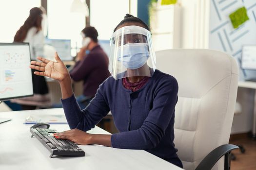 Pov of african entrepreneur waving on video call with business people weaing face mask against covid19. Woman speaking with team during online conference while colleagues working in background.