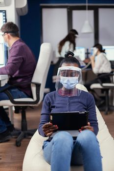 African manager taking notes on tablet pc wearing face mask against covid19 at workplace. Multiethnic business team working respecting social distance during global pandemic with coronavirus.