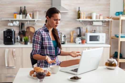 Stresed woman using laptop in kitchen during breakfast with cup of green tea. Working from home using device with internet technology, browsing, searching on gadget in the morning.