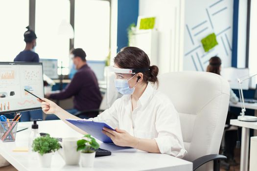 Manager wearing protection against covid 19 during global pandemic. Employees with visors working in corporate company workspace respecting social distance analysing data and graphs.