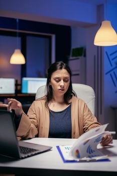 Focused entrepreneur looking at charts trying to finish deadline in empty office. Woman checking notes on clipboard while working on important project doing overtime.