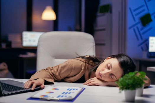 Tired businesswoman resting head on desk holding hand on laptop. Employee falling asleep while working late at night alone in the office for important company project.