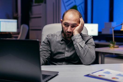 Tired businessman sit at the computerin the evening works on deadline. Workaholic employee falling asleep because of working late at night alone in the office for important company project.