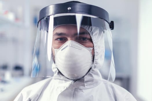 Tired healthcare scientist loooking at camera wearing ppe suit with face shieldin lab. Overworked researcher dressed in protective suit against invection with coronavirus during global epidemic.