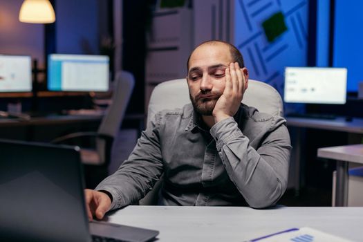 Bored sleepy businessman working on laptop doing overtime at workplace. Workaholic employee falling asleep because of working late at night alone in the office for important company project.