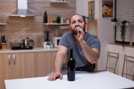 Pensive man drinking glass of wine while sitting alone and depressed. Person with alcohol addiction having bottle of alcoholic beverage, liquor, booze, feeling heartbroken and desperate