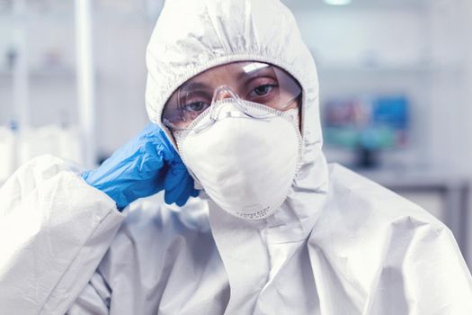 Portrait of overworked biolog looking at camera in modern equipped lab. Tired woman scientist in biotechnology laboratory wearing protective suit during global epidemic.