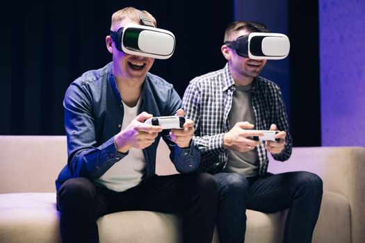 Overjoyed men friends winning videogame having fun together on sofa celebrating victory at home. Excited funny young gamers wear virtual reality glasses holding controllers playing video game.