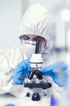 Medical engineer in ppe suit in modern laboratory looking at coronavirus sample using microscope. Chemist researcher during global pandemic with covid-19 checking sample in biochemistry lab