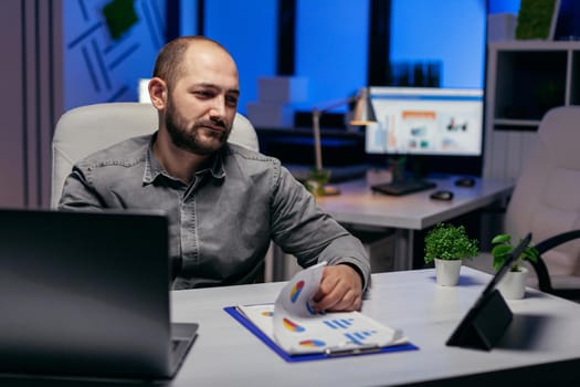 Businessman working late at night in workplace using tablet pc holding hand on clipboard with chart. Bearded entrepreneur doing overhourse at the office in the evening to finish a deadline.