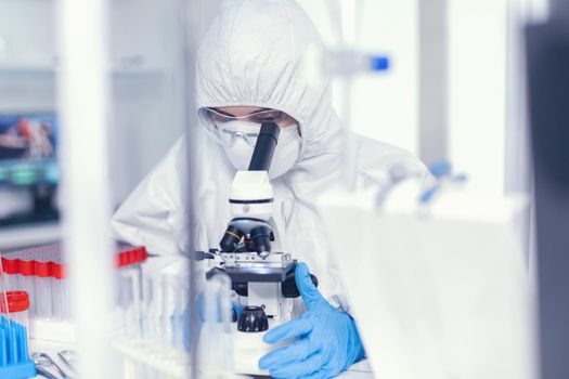 Scientist in sterile protection suit analysing coronavirus looking through microscope. Chemist in coverall examining vaccine evolution using high tech researching diagnosis against covid19 virus