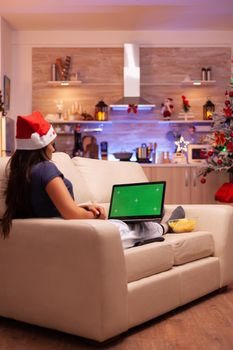 Female person resting on sofa in xmas decorated kitchen holding green screen mock up chroma key laptop computer with isolated display. Adult woman enjoying christmas holiday celebrating winter season