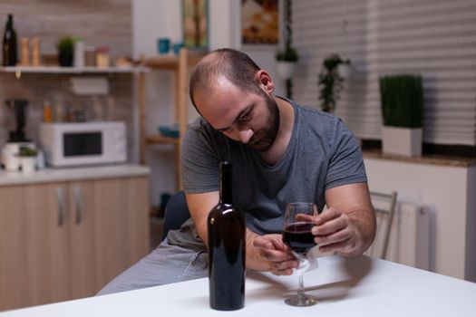 Caucasian man holding glass of wine sitting in kitchen at home. Depressed person drinking liquor, booze, alcoholic beverage alone while feeling intoxicated with bottle of alcohol