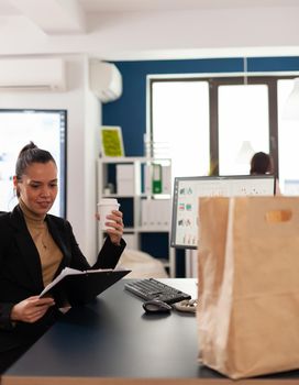 Businesswoman sitting at desk in corporate office reading financial statistics on clipboard, before enjoying tasty delicous food from takeaway in paper bag.