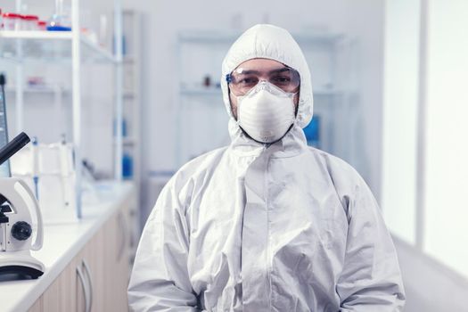 Portrait of tired scientist wearing coverall with protection glasses looking at camera in medicine laboratory. Overworked researcher dressed in protective suit against invection with coronavirus during global epidemic.