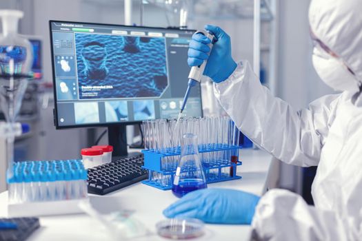 Medic assistant working in biochemistry lab working with automatic pipette wearing protection suit. Chemist in modern laboratory doing research using dispenser during global epidemic with covid-19.