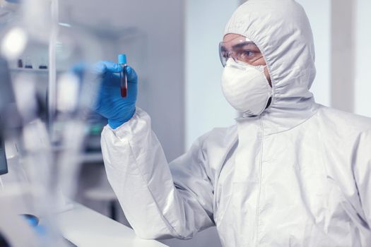 Medic during coronavirus outbreak dressed in biohazd suit holding blood in tube. Doctor working with various bacteria and tissue, pharmaceutical research for antibiotics against covid19.