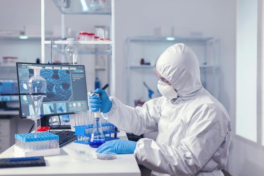 Man taking sample of liquid from glass flask using micropipette dressed in protection suit. Chemist in modern laboratory doing research using dispenser during global epidemic with covid-19.