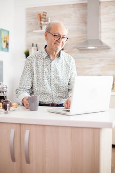 Elderly man smiling while holding a mug of coffee working on laptop in his kitchen. Daily life of Senior man working on laptop in kitchen during breakfast holding a cup of coffee. Elderly retired person working from home, telecommuting using remote internet job online communication on modern technology notebook