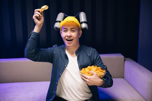 Young male sits on a sofa and eats snacks while watching a movie. Handsome man with beer helmet on the head watching TV and eating potato chips on the couch at home.