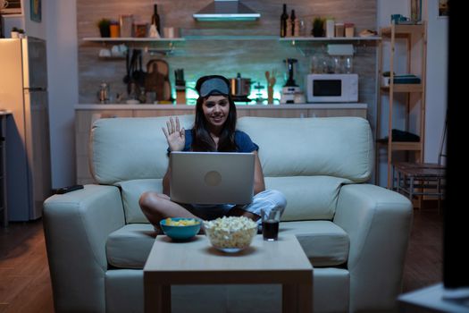 Lady with eye mask having video calling on laptop computer at night. Exhausted person in pijamas talking on notebook computer webcam with colleagues sitting on couch at home using internet technology