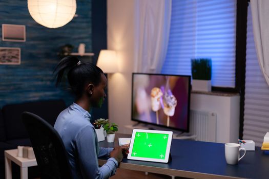 Black entrepreneur woman working on project deadline from home at night in home office holding tablet pc with green screen. Using mockup chroma key display computer.