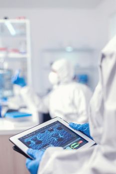 Chemist dressed in protection suit for coronavirus holding tablet pc in laboratory. Team of scientists conducting vaccine development using high tech technology for researching treatment against covid19