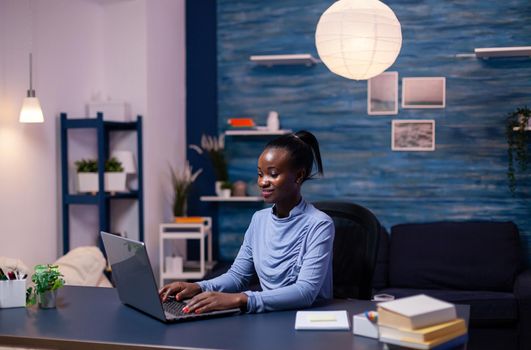 African woman working remotly from home office in the evening on laptop computer. Black entrepreneur sitting in personal workplace writing on keyboard.