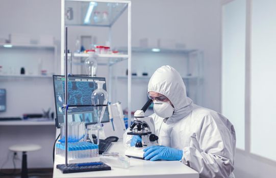 Scientist studying new virus in laborator using microscope dressed in protection suit. Virolog in coverall during coronavirus outbreak conducting healthcare scientific analysis.