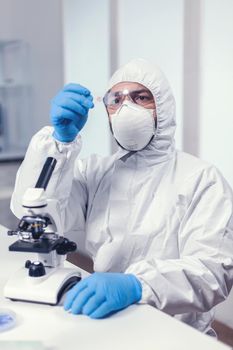 Medical engineer looking attentive at sample on glass slide sitting at his workplace dressed in ppe suit. Examining vaccine evolution using high tech for researching treatment against covid19 virus.