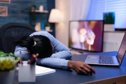 Exhausted dark skinned woman resting on office desk in home office late at night. Busy employee using modern technology network wireless doing overtime sleeping on table.