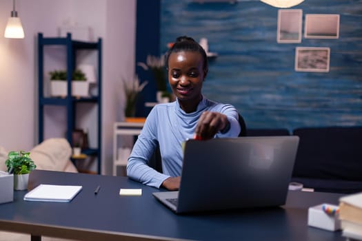 Creative african woman placing sticky notes on laptop working in the evening to finsih a project for job. Black freelancer respecting deadline studying late at night.