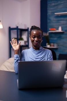 African woman waving at laptop webcam in the course of video conference working late at night from home office. Black freelancer working with remotely team chatting virtual online conference.