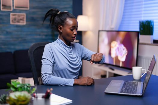 Worried african entrepreneur looking at wrist watch working hard to finish a big project for work. Nervous black manager doing overtime working on computer.