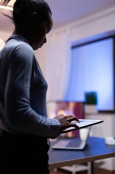 Dark skinned woman utilises tablet pc in home office late at night. Busy focused employee using modern technology network wireless doing overtime writing.