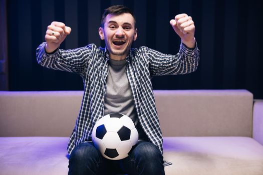 Handsome Caucasian man worried and cheering up for football game on TV sitting on sofa late at night. Close up of man with ball in hands watch sport channel.