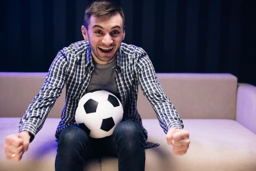 Sports Fan Reaction Concept. Fun guy 30s football fan cheer up support favorite team hold soccer ball in t-shirt in dark living room. People emotions sport leisure lifestyle.