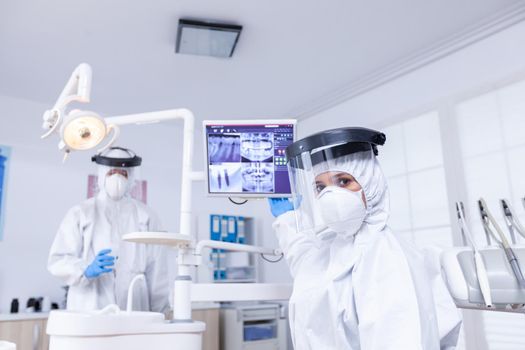 Doctor showing to X Ray on dental unit. Stomatology specialist wearing protective suit against infection with coronavirus pointing at radiography.