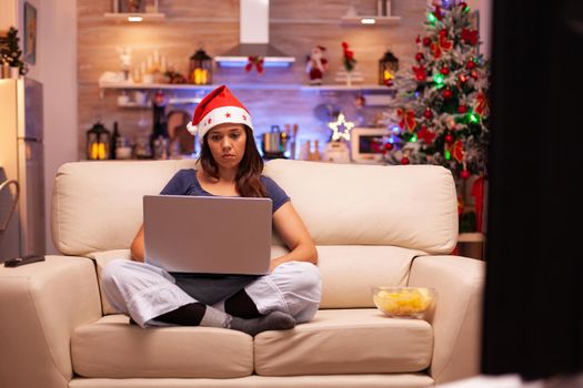 Woman with santa hat sitting on sofa checking business email using laptop computer searching information using laptop computer. Girl spending christmas time working. Winter season