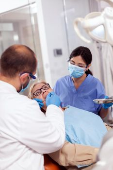 Dentist with assistant install implant in patient mouthin modern clinic. Elderly woman during medical examination with stomatolog in dental office with orange equipment.