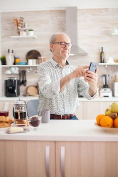 Retired old person scrolling on the phone, using internet and smartphone in kitchen during breakfast. Elderly person using internet online chat technology video webcam making a video call connection camera communication conference call