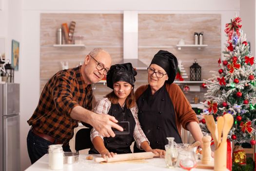 Grandparent and child taking selfie on christmas day wearingapron and bonette. Happy cheerful joyfull teenage girl helping senior woman preparing sweet cookies to celebrate winter holidays.