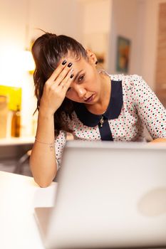 Businesswoman looking exhausted and massaging forhead while working overtime on laptop from home kitchen. Employee using modern technology at midnight doing overtime for job, business, career, network, lifestyle