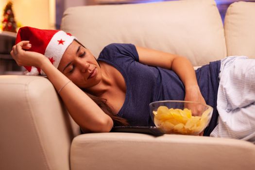 Girl falling asleep on couch in xmas decorated kitchen after watching entertainment movie on television. Caucasian female with red santa hat celebrating christmas holiday. X-mas season
