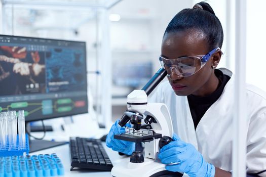 Medical researcher with african ethnicity using microscope doing investigation and evaluates sample. Black healthcare scientist in biochemistry laboratory wearing sterile equipment.