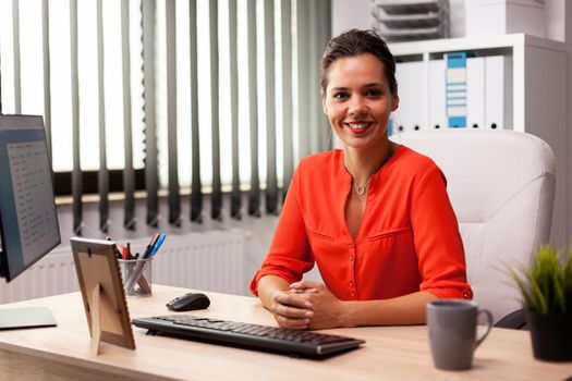 Executive freelancer finance entreprenerur wearing red blouse smiling at camera in workplace. Successful confident woman in marketing sitting at desk in workplace using computer.