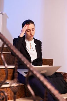 Exhausted worried businesswoman using laptop to finish corporate project deadline. Serious entrepreneur working on job project sitting on staircase of business building at night hours for job.