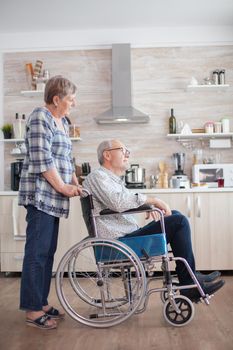 In the kithcen old woman is looking at handicapped husband who is in wheelchair. Disabled senior man sitting in wheelchair in kitchen looking through window. Living with handicapped person. Wife helping husband with disability. Elderly couple with happy marriage.