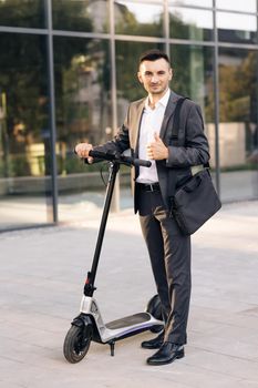 Young businessman in elegant suit standing outdoors, cheerfully smiling and showing thumb up. Handsome businessman with his electric scooter in front the business building.