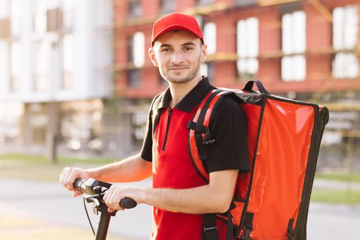 Portrait shot of man delivery worker in red uniform standing at street and turning face to camera. Male courier with red backpack and electric scooter. Delivering green transportation.